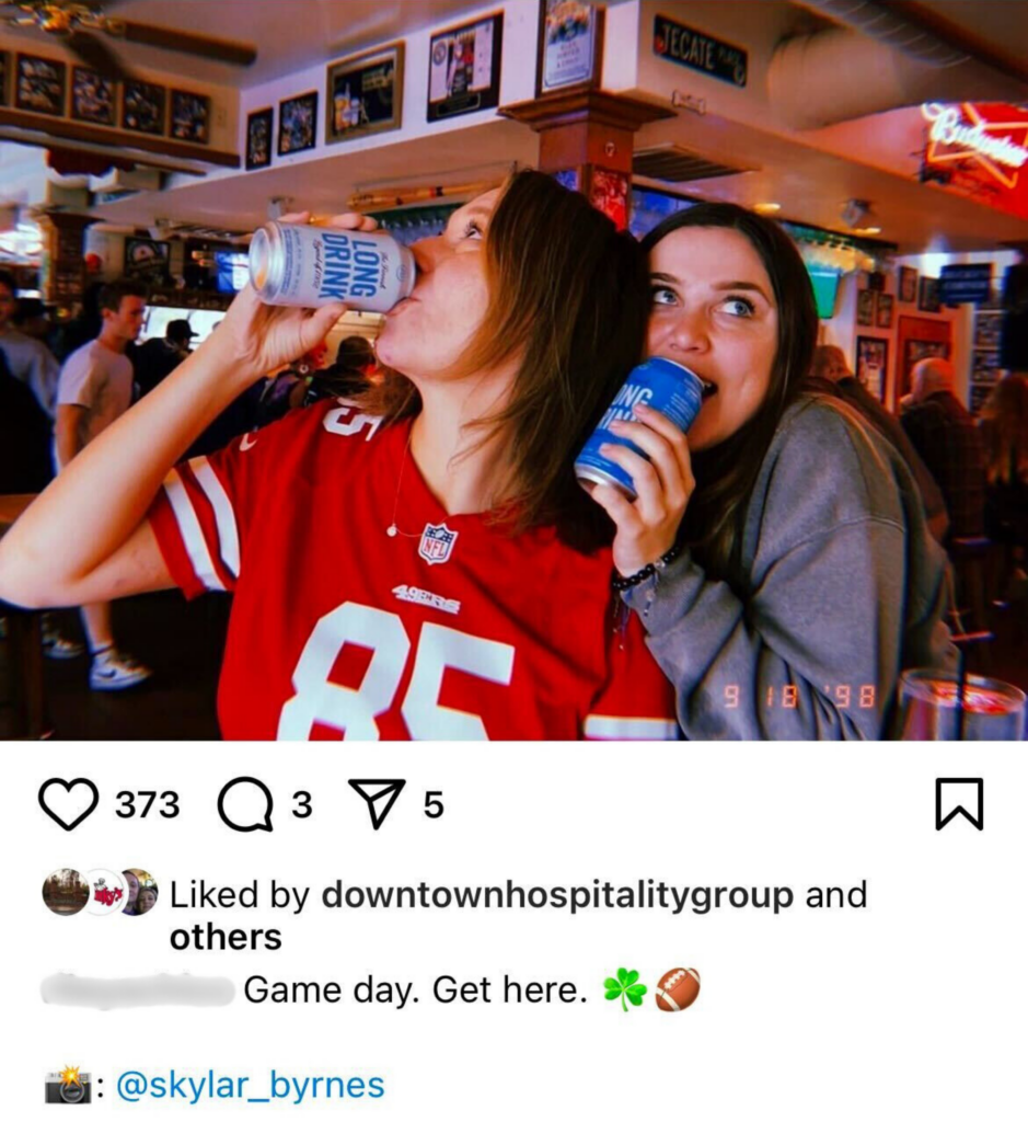 Two friends enjoying canned drinks at a bar, one wearing a 49ers jersey and the other in casual attire, surrounded by sports decor and a lively bar atmosphere.